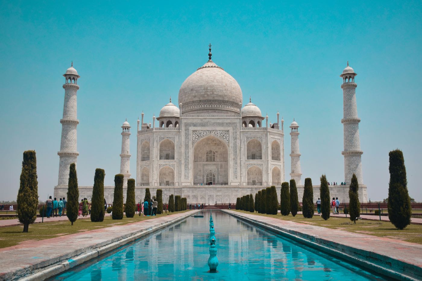 The Taj Mahal seen straight down the reflecting pool, its dome and four minarets in white marble under a clear sky.