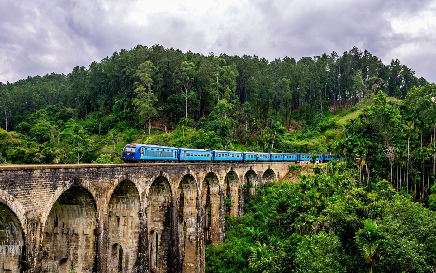 A blue train crossing the stone arches of Nine Arch Bridge at Ella, surrounded by dense green hillside forest.
