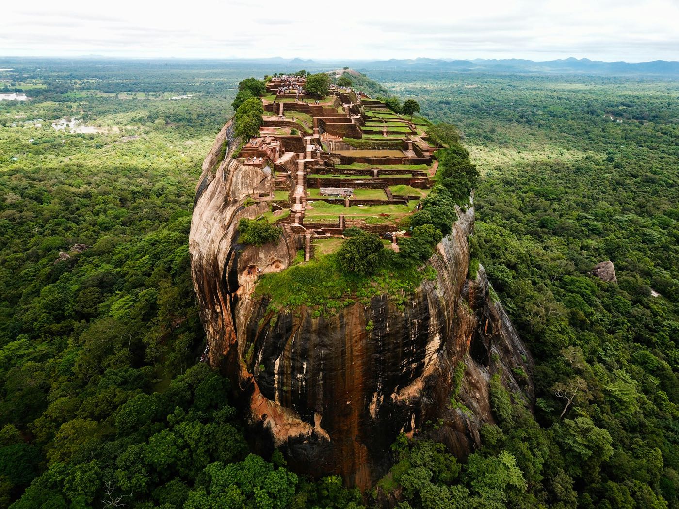 Sigiriya rock from the air: a sheer flat-topped outcrop rising from unbroken green jungle, its summit covered in the terraces and brick foundations of a ruined palace.